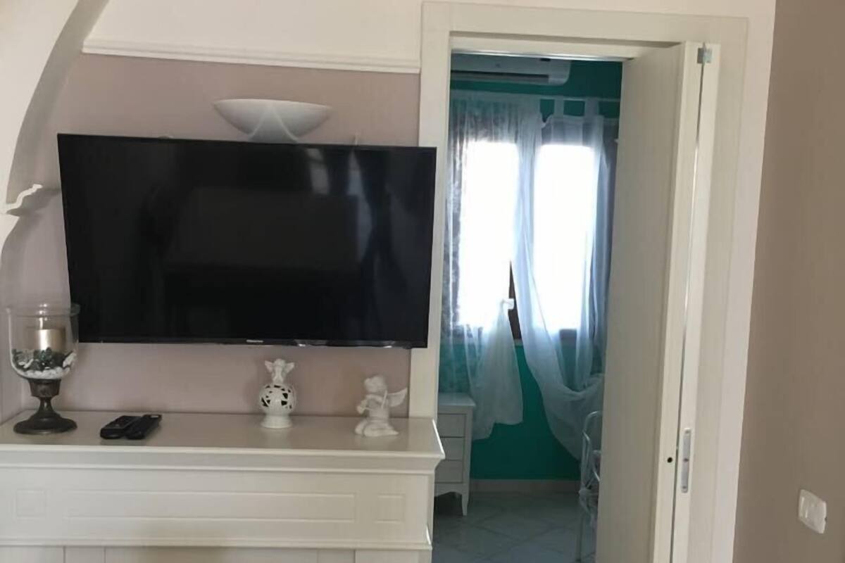 A room with a TV on a white cabinet, a small table with a glass and figurines, and a door leading to another room with teal walls.