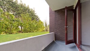 Un balcón con vista a un área verde y árboles, un muro de ladrillo y una puerta entreabierta.