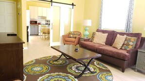 A living room with a sofa, coffee table, and patterned rug. There is a kitchen in the background with cabinets and a dining area. - Keep Cool Apartment (Gros Islet)
