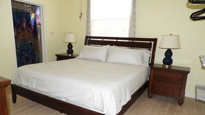 A bedroom with a wooden bed, two bedside tables with lamps, a window with curtains, and a patterned wall hanging. - Keep Cool Apartment (Gros Islet)