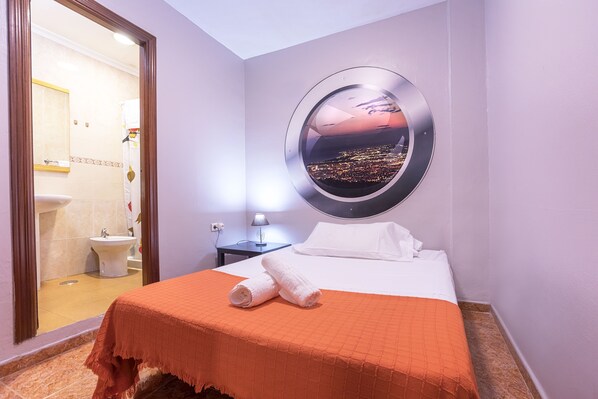 A hotel room with a bed, a round mirror with a cityscape view, a bathroom with a toilet and sink, and a nightstand with a lamp. - Hostal Ciudad Jardin (Málaga)