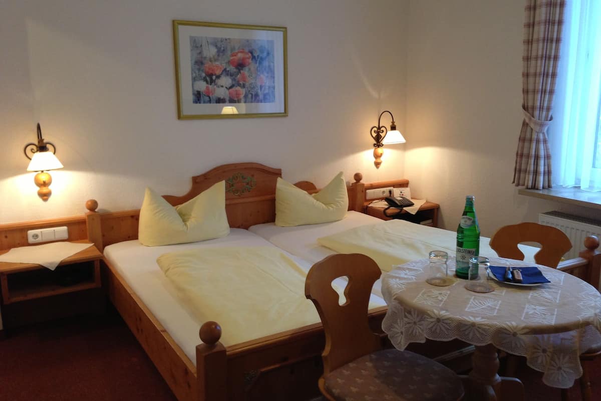 A hotel room with two beds, a small table with a tablecloth, and a chair. There is a painting on the wall, a lamp on each side of the bed, and a bottle of water on the table.