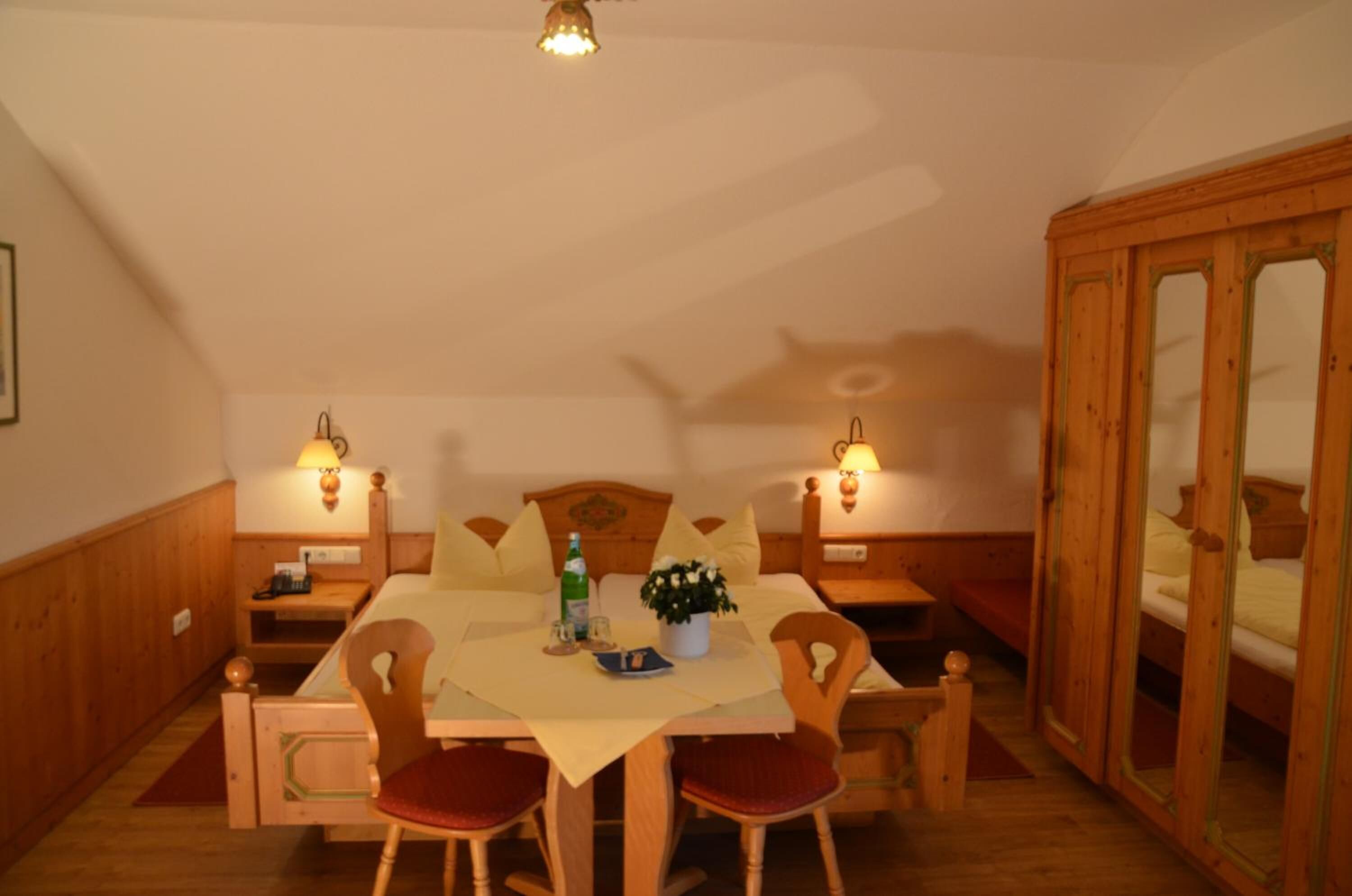 A cozy dining area with a wooden table, chairs, and a bench. There are two wall-mounted lamps, a small table with a bottle and glasses, and a potted plant on the table.