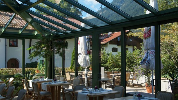 An outdoor dining area with tables and chairs, a glass roof, and a mountain view. - Gästehaus Maria (Garmisch-Partenkirchen)
