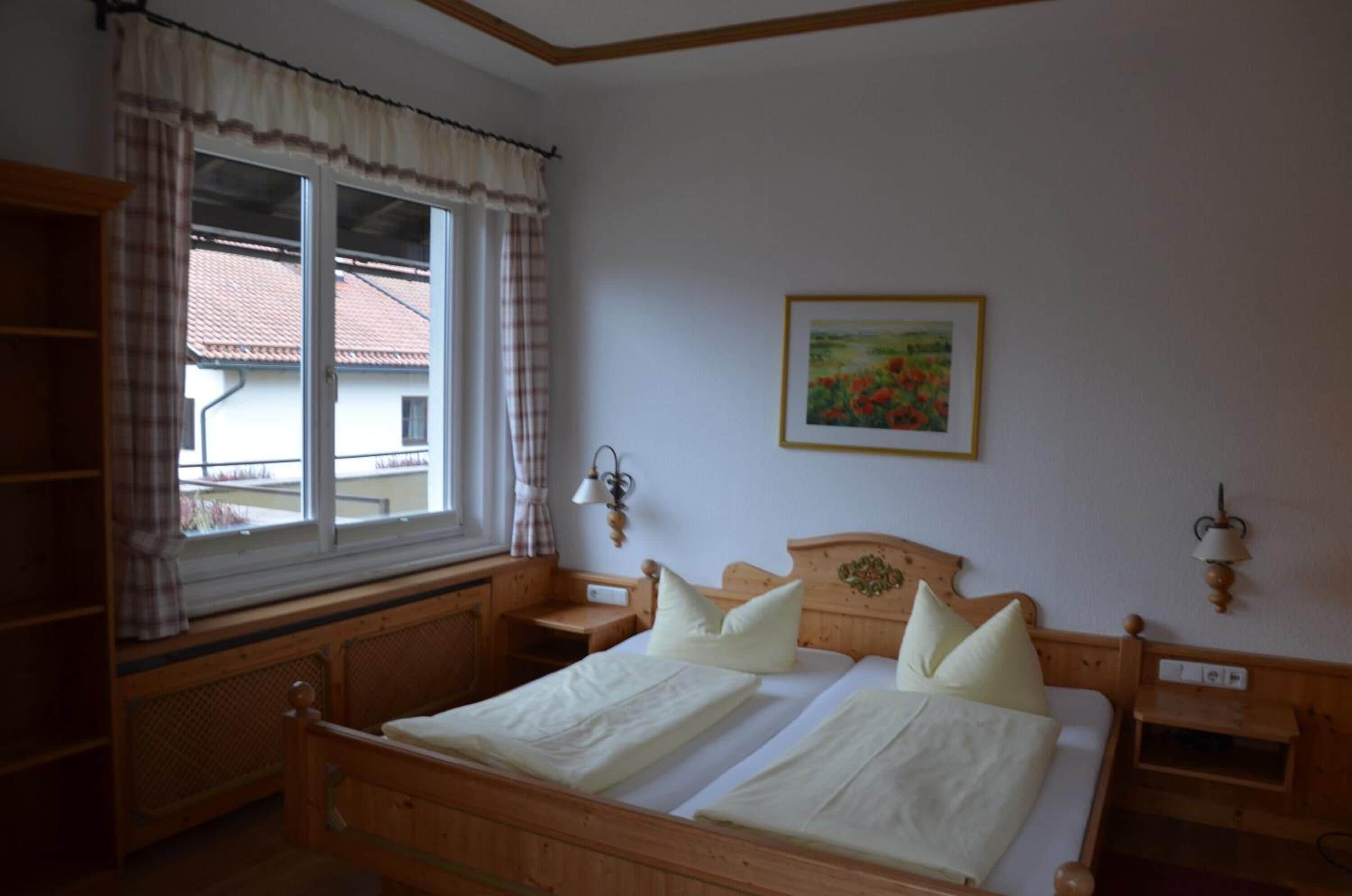 A bedroom with a wooden bed, a window with curtains, a painting on the wall, and a nightstand with a lamp.