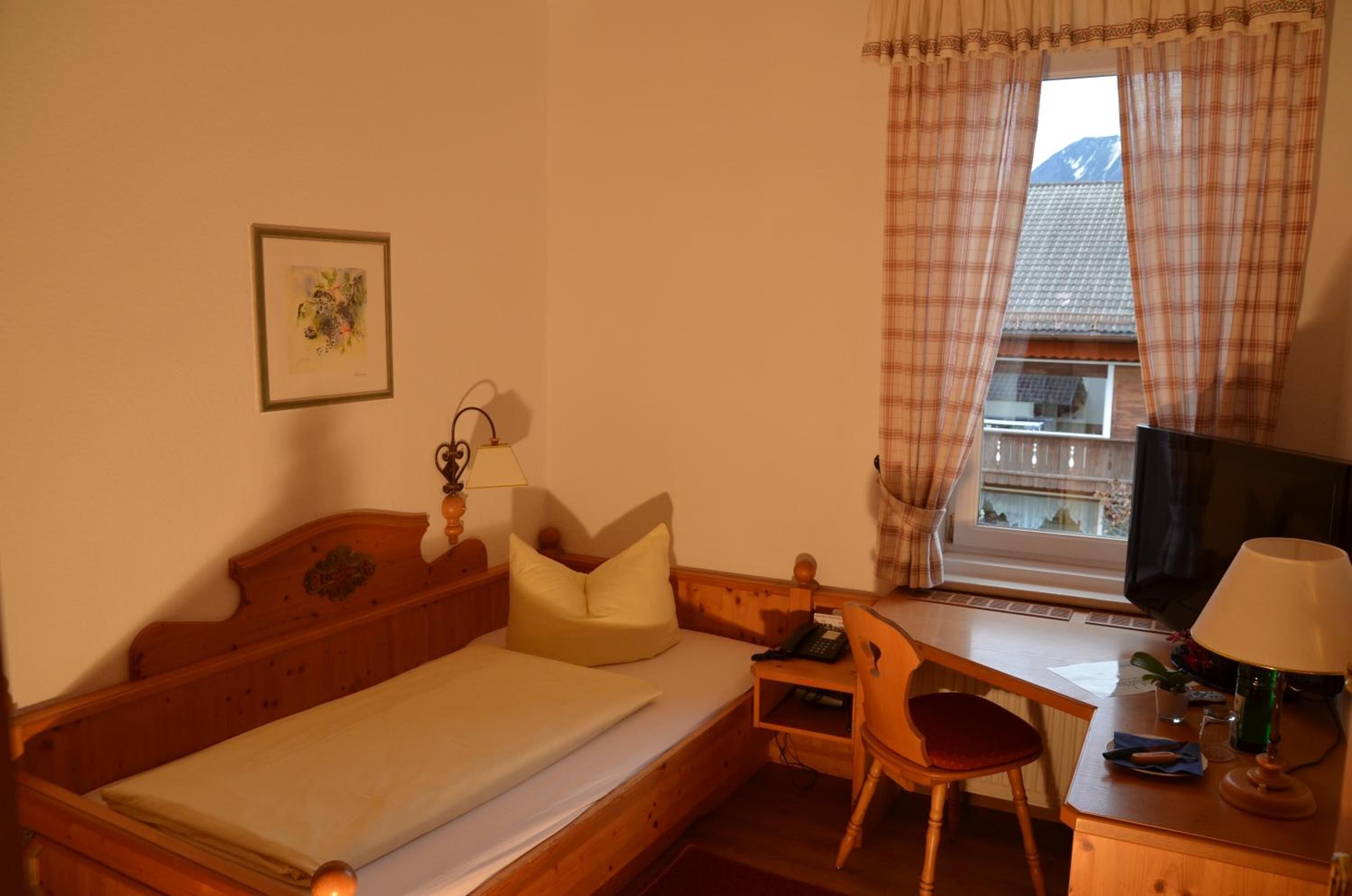 Um quarto pequeno e acolhedor com uma cama de madeira, uma escrivaninha, uma cadeira, uma televisão e uma janela com cortinas.