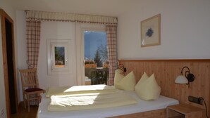 A bedroom with a wooden bed, a chair, a bedside table, and a window with curtains. - Gästehaus Maria (Garmisch-Partenkirchen)