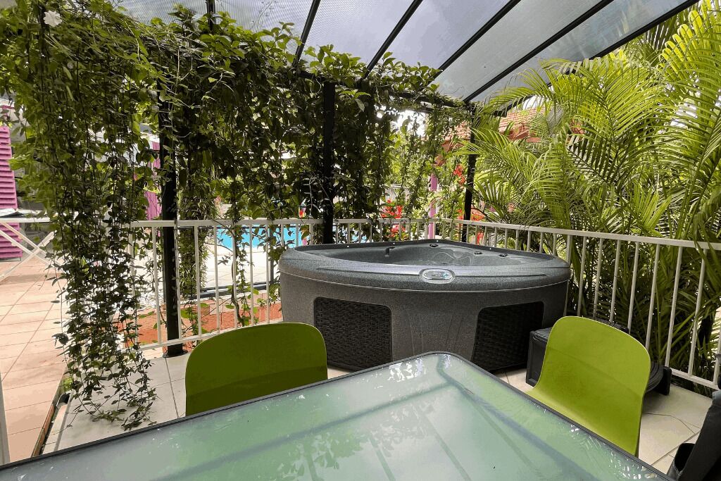 An outdoor seating area with a hot tub, surrounded by lush greenery and a glass table.
