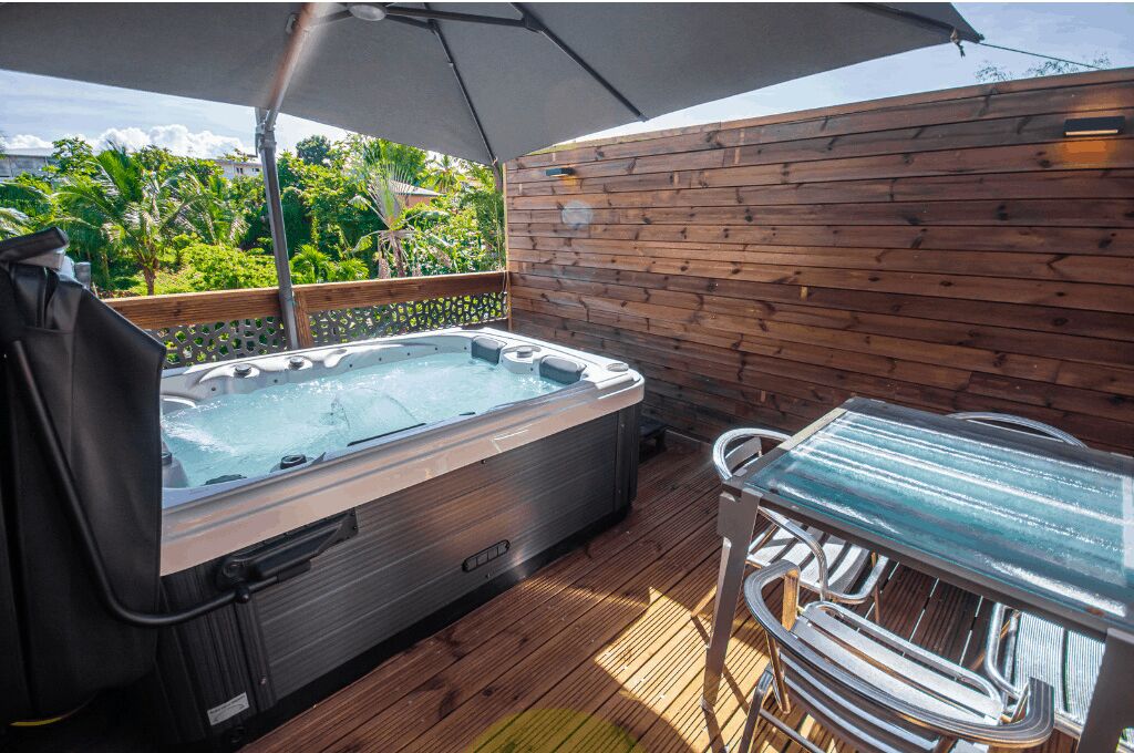 Un jacuzzi en una terraza de madera con una mesa y sillas, rodeado por una cerca de madera y abundante vegetación.