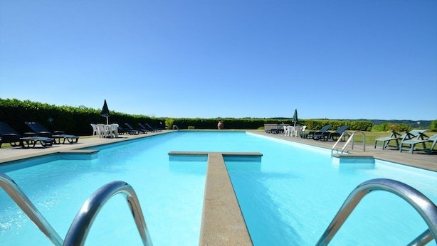 A clear blue swimming pool with a central diving board, surrounded by lounge chairs and umbrellas.