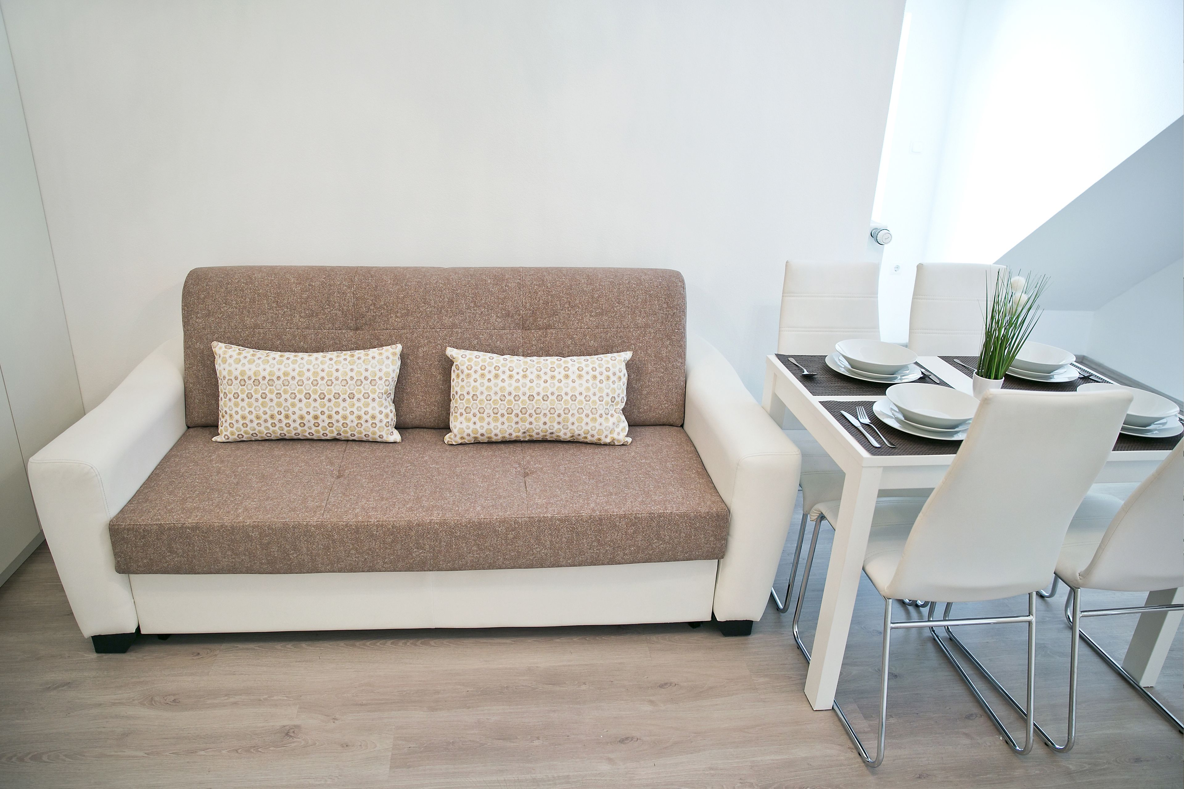 Una sala de estar moderna con un sofá marrón, dos cojines estampados, una mesa de comedor con sillas blancas y un jarrón con plantas verdes.