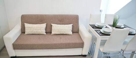 A modern living room with a brown sofa, two patterned pillows, a dining table with white chairs, and a vase with green plants.