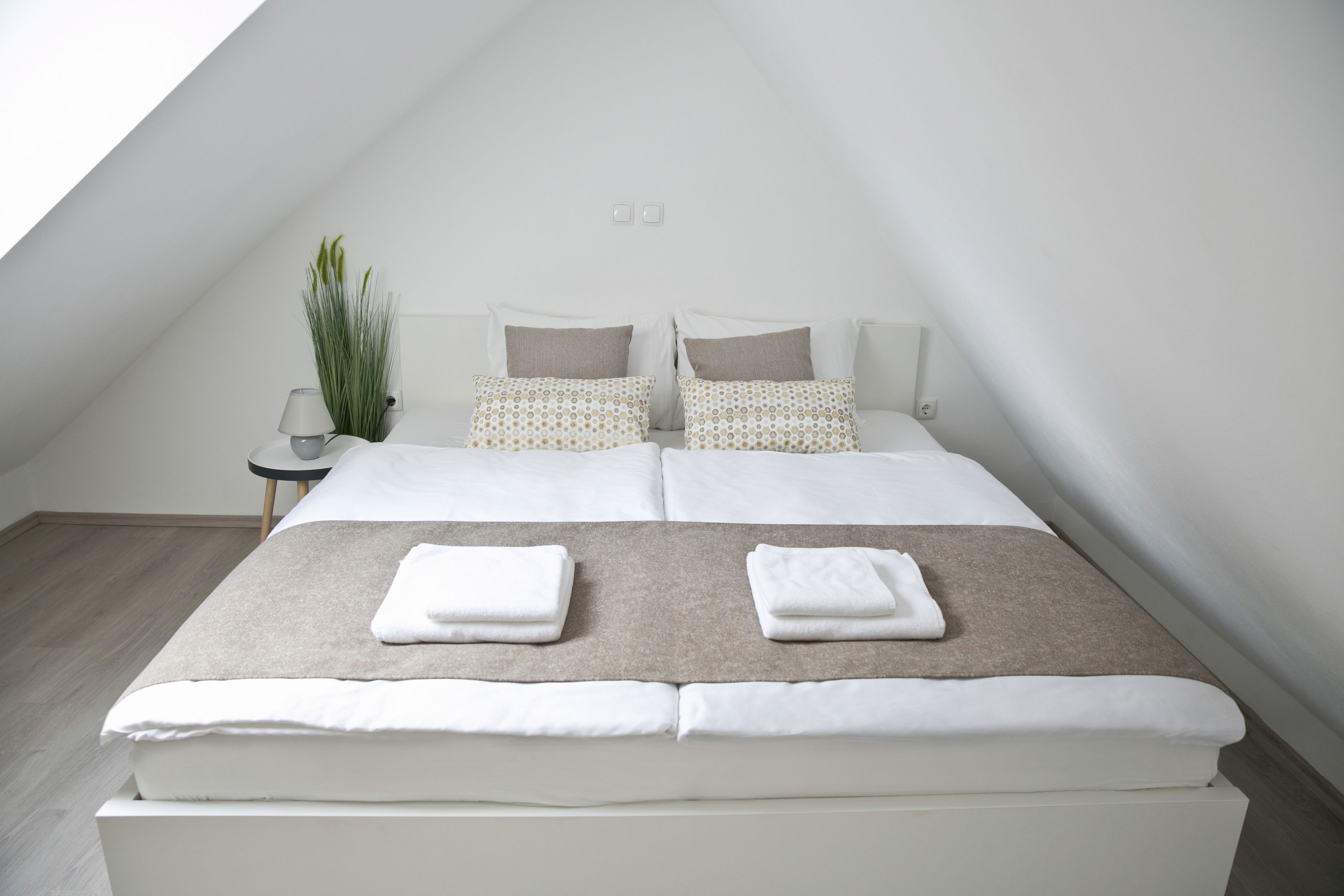 Ein modernes Schlafzimmer mit schrägen Wänden, einem großen Bett mit weißer und beiger Bettwäsche, zwei Nachttischen und einem Blumentopf.
