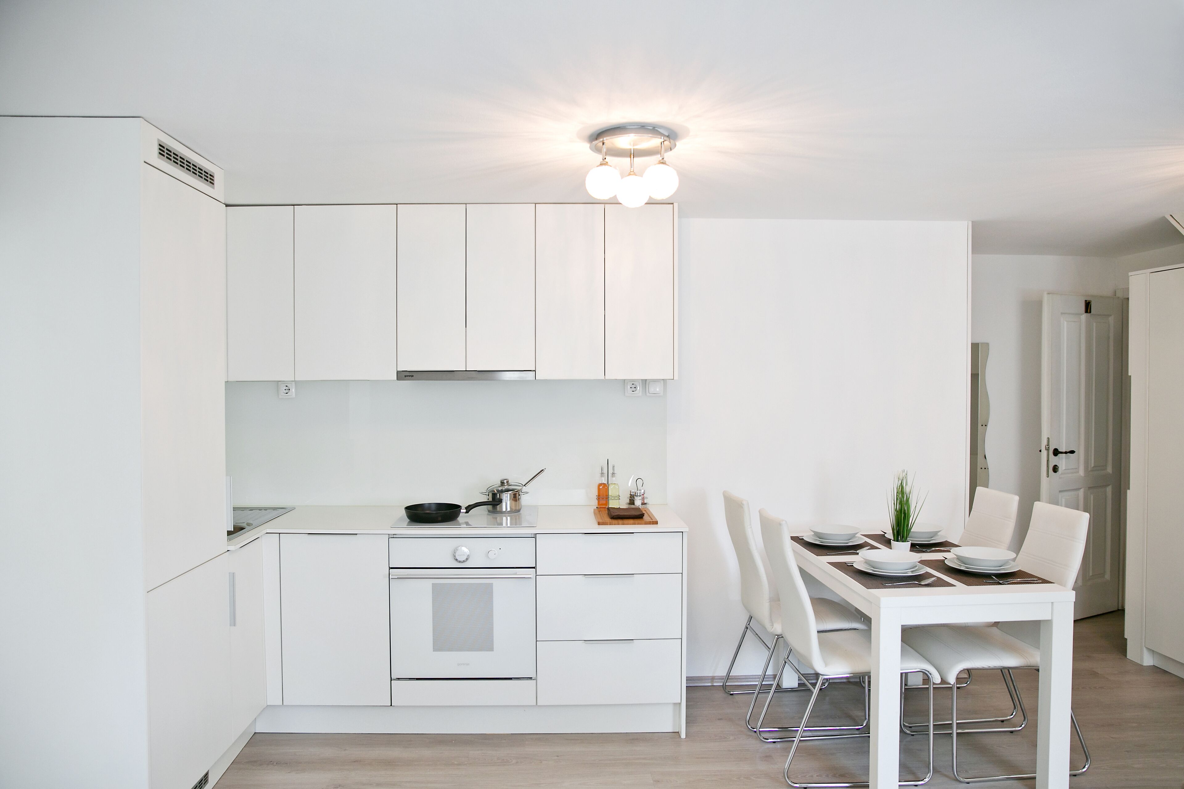 Una cocina moderna con armarios blancos, horno empotrado y comedor con mesa y sillas blancas.
