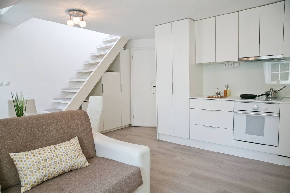 Eine moderne Küche mit weißen Schränken, einem eingebauten Ofen und einem Essbereich mit einer braunen Couch und gemusterten Kissen.