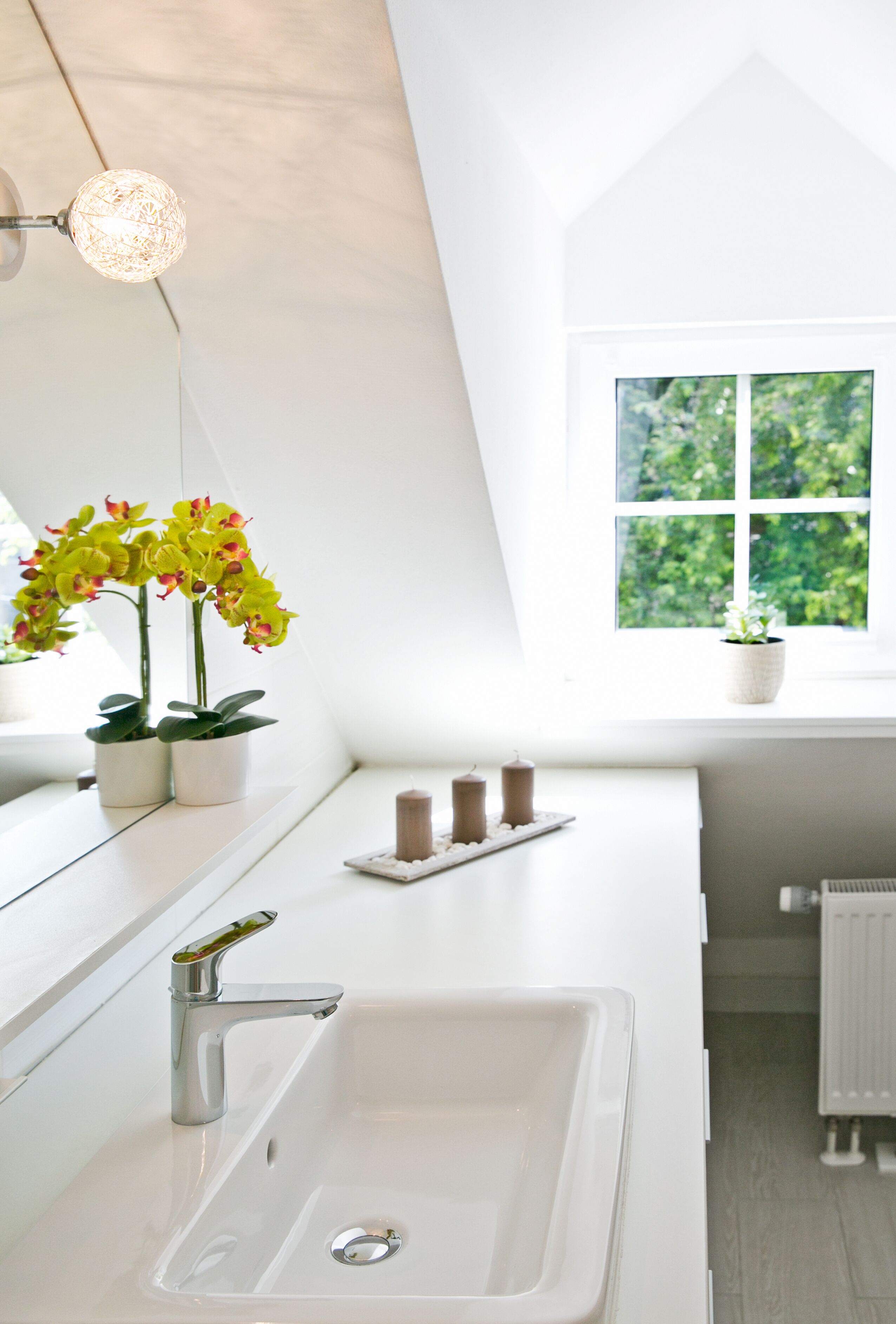 Un baño moderno con un lavamanos blanco, un espejo, una ventana con vistas a la vegetación y dos orquídeas en macetas.