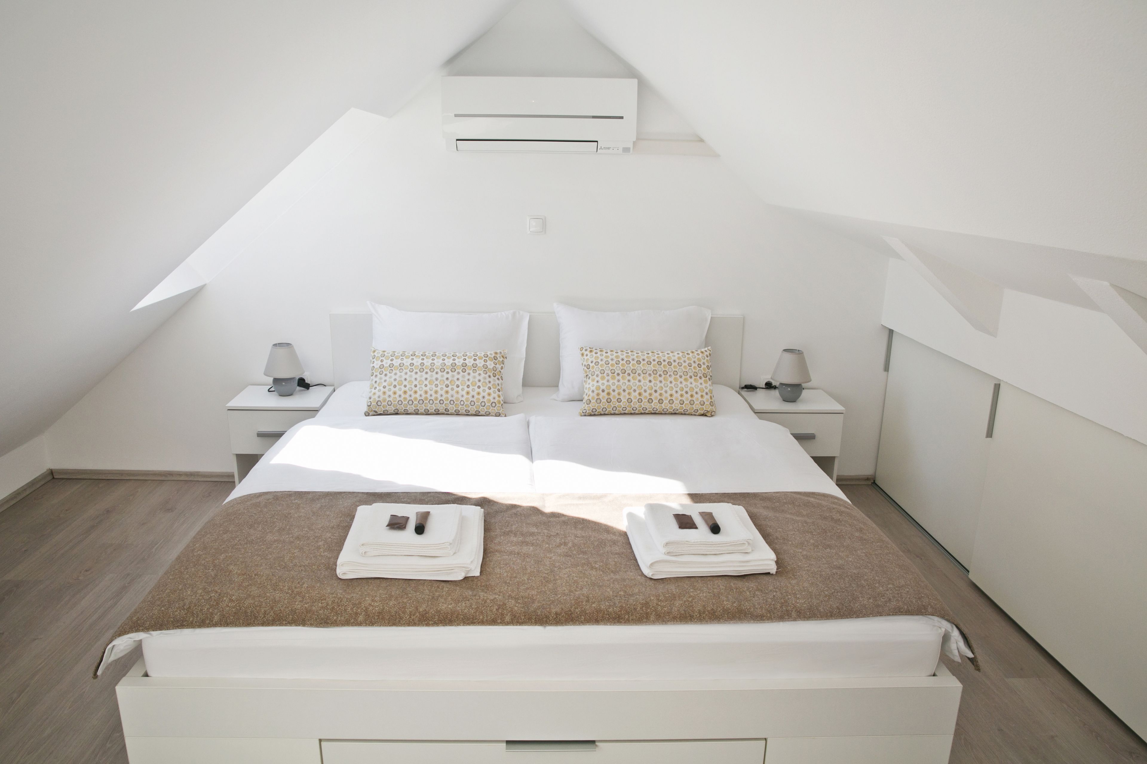 A modern bedroom with a sloped ceiling, a central bed with two pillows, bedside tables with lamps, and a neatly made bed with folded towels.