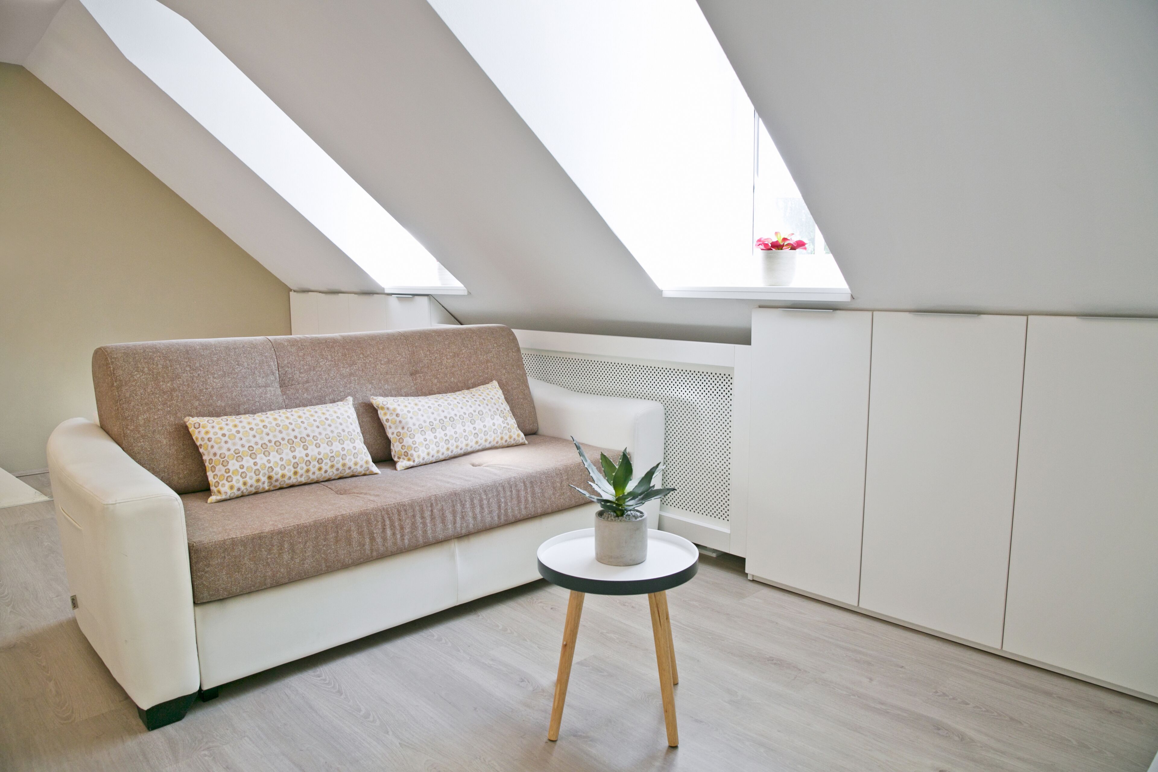 A modern loft apartment with a sloped ceiling, a white sofa with patterned cushions, a small round table with a plant, and a radiator.