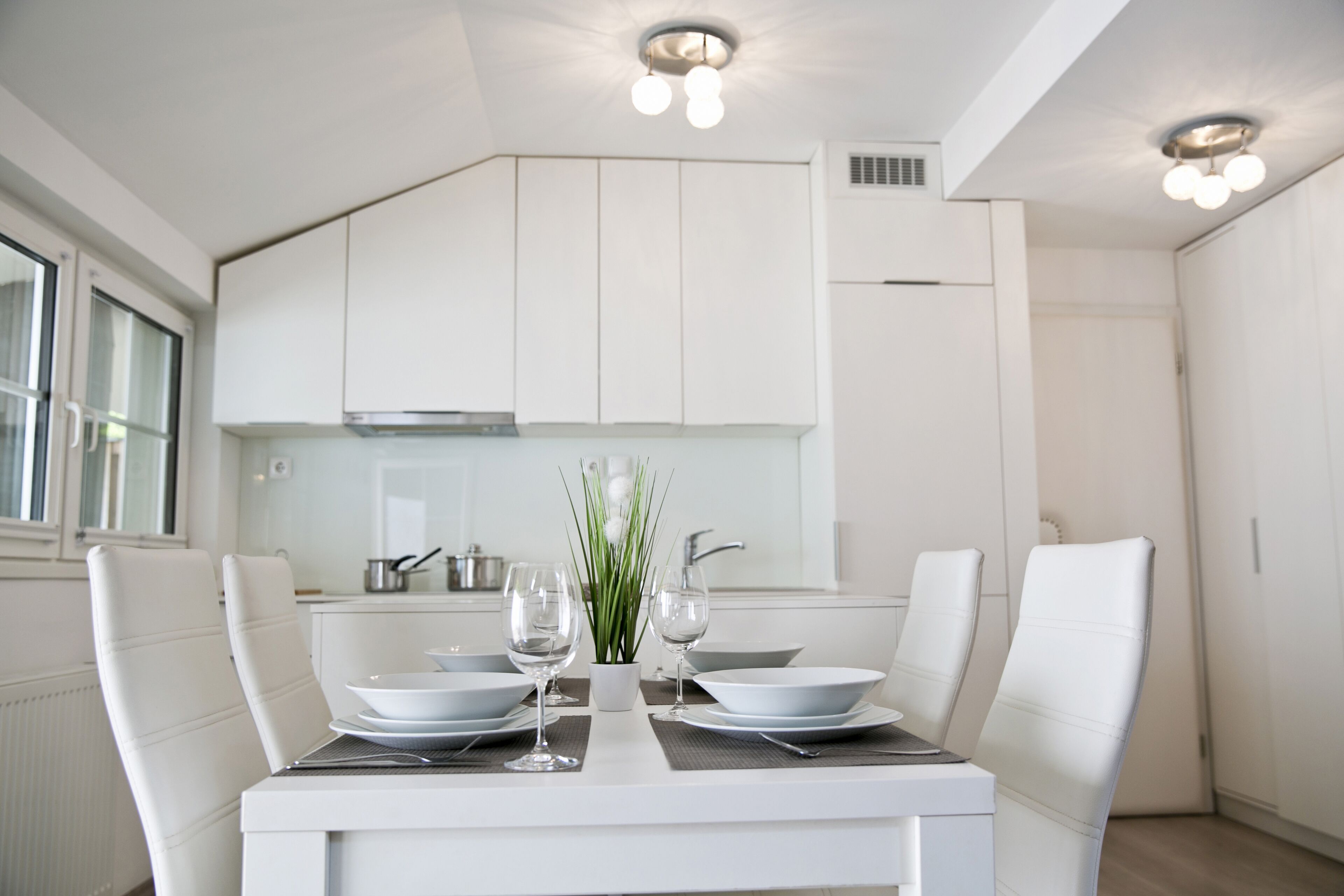 Une cuisine moderne avec des meubles blancs, une table à manger dressée pour deux personnes et une plante en pot posée sur la table.