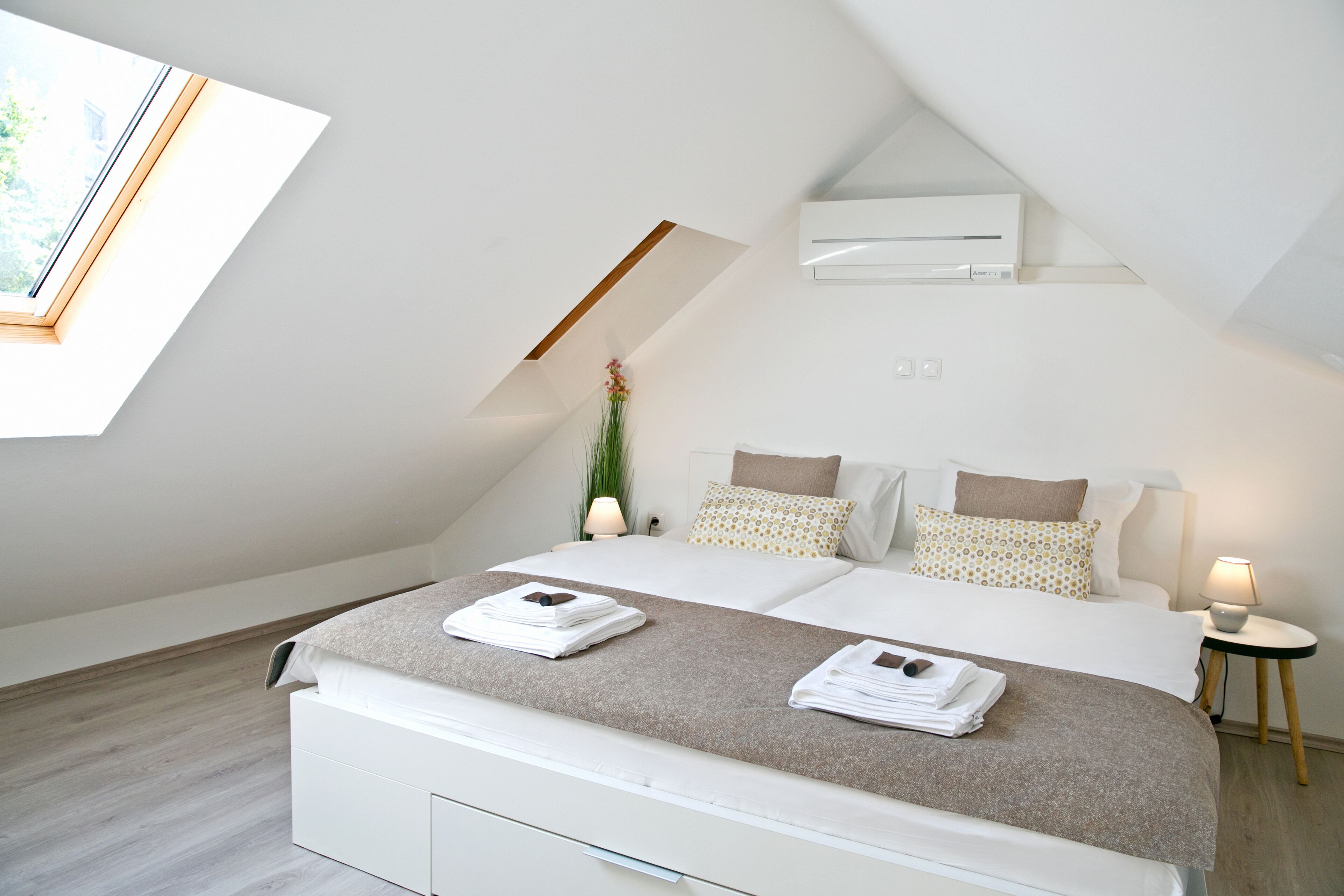 Une chambre moderne avec une lucarne, un lit recouvert de linge de lit blanc et agrémenté d’oreillers à motifs, une table de chevet avec une lampe et une petite plante en pot.