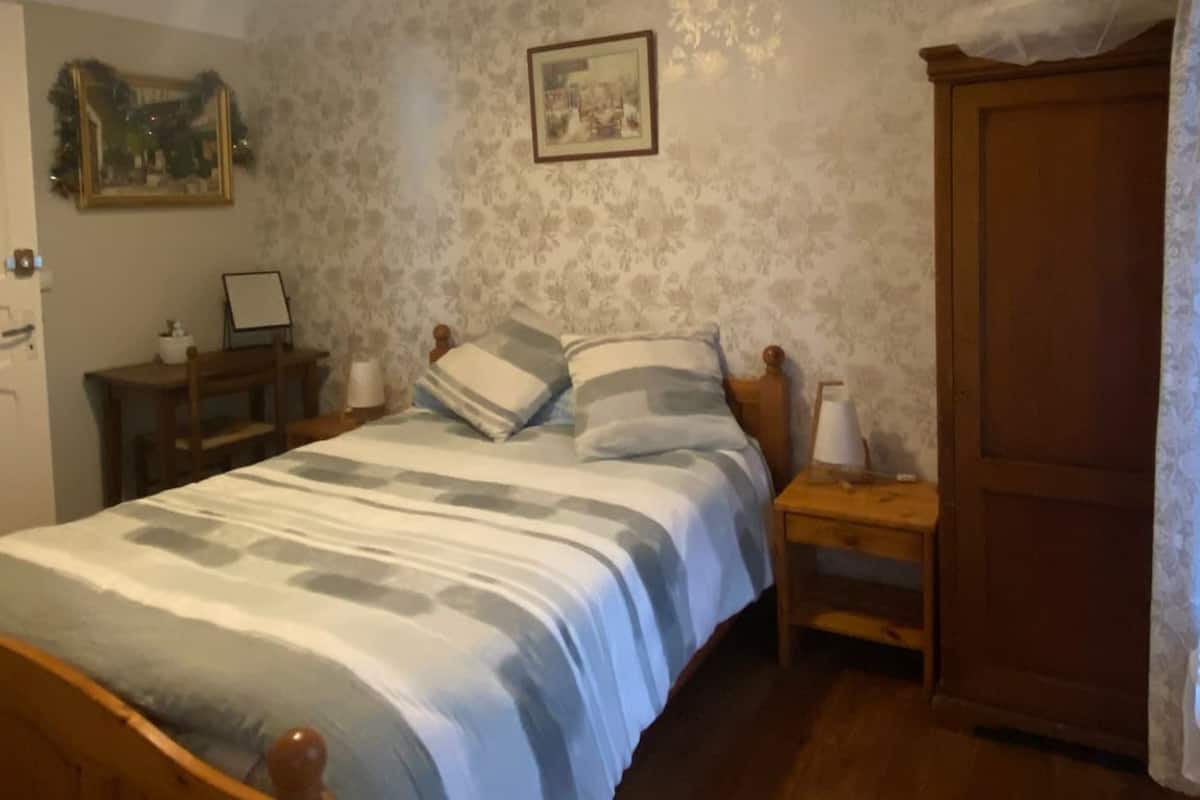 A bedroom with a wooden bed, a nightstand, a desk with a chair, a wardrobe, and a framed picture on the wall.