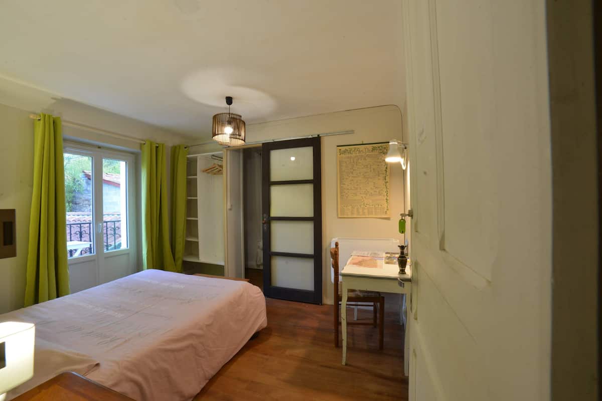 A bedroom with a bed, a desk, a chair, a window with curtains, and a door leading to another room.
