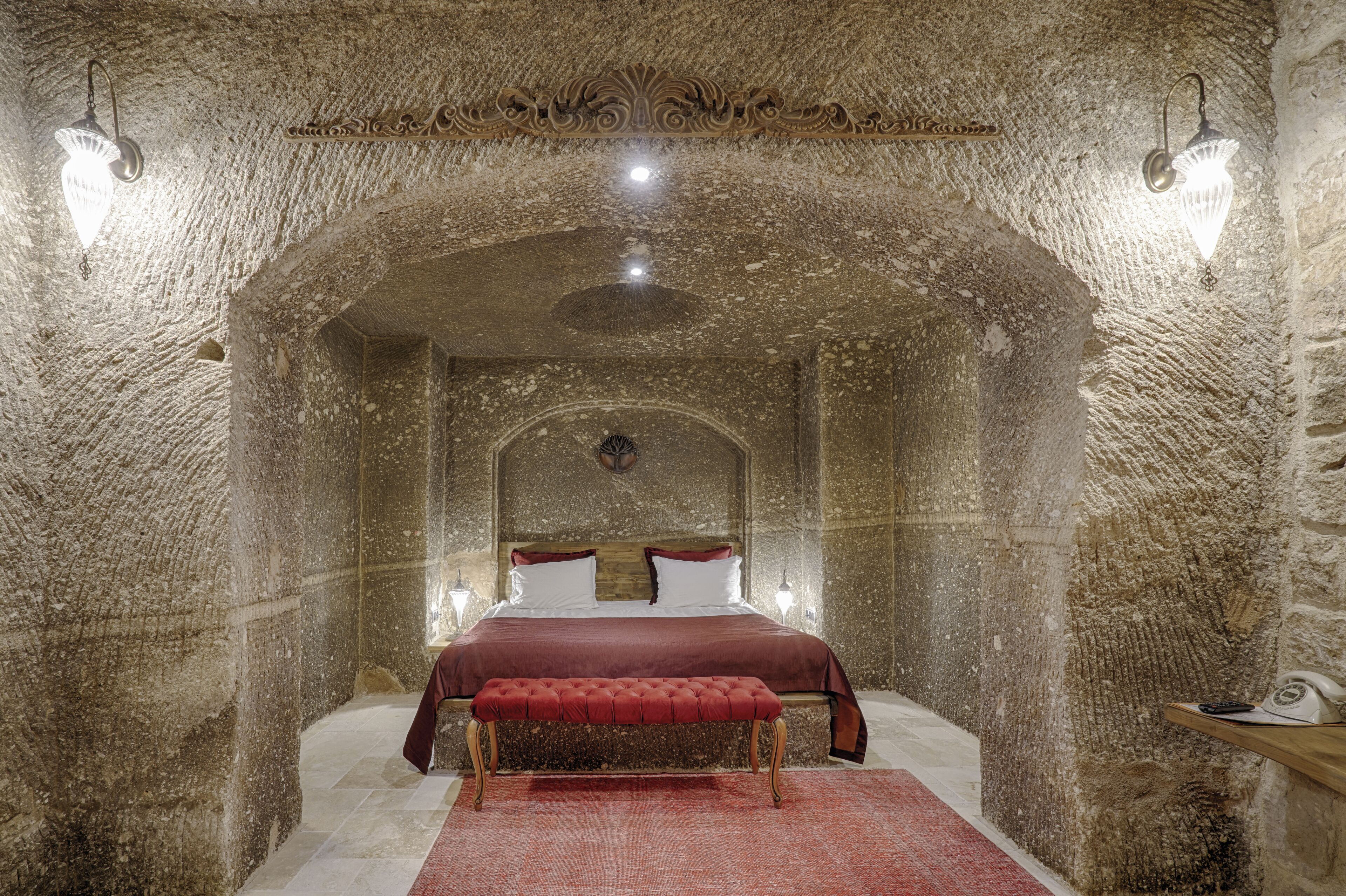 A bedroom with a stone archway, a bed with a red bedspread, and two wall-mounted lights.