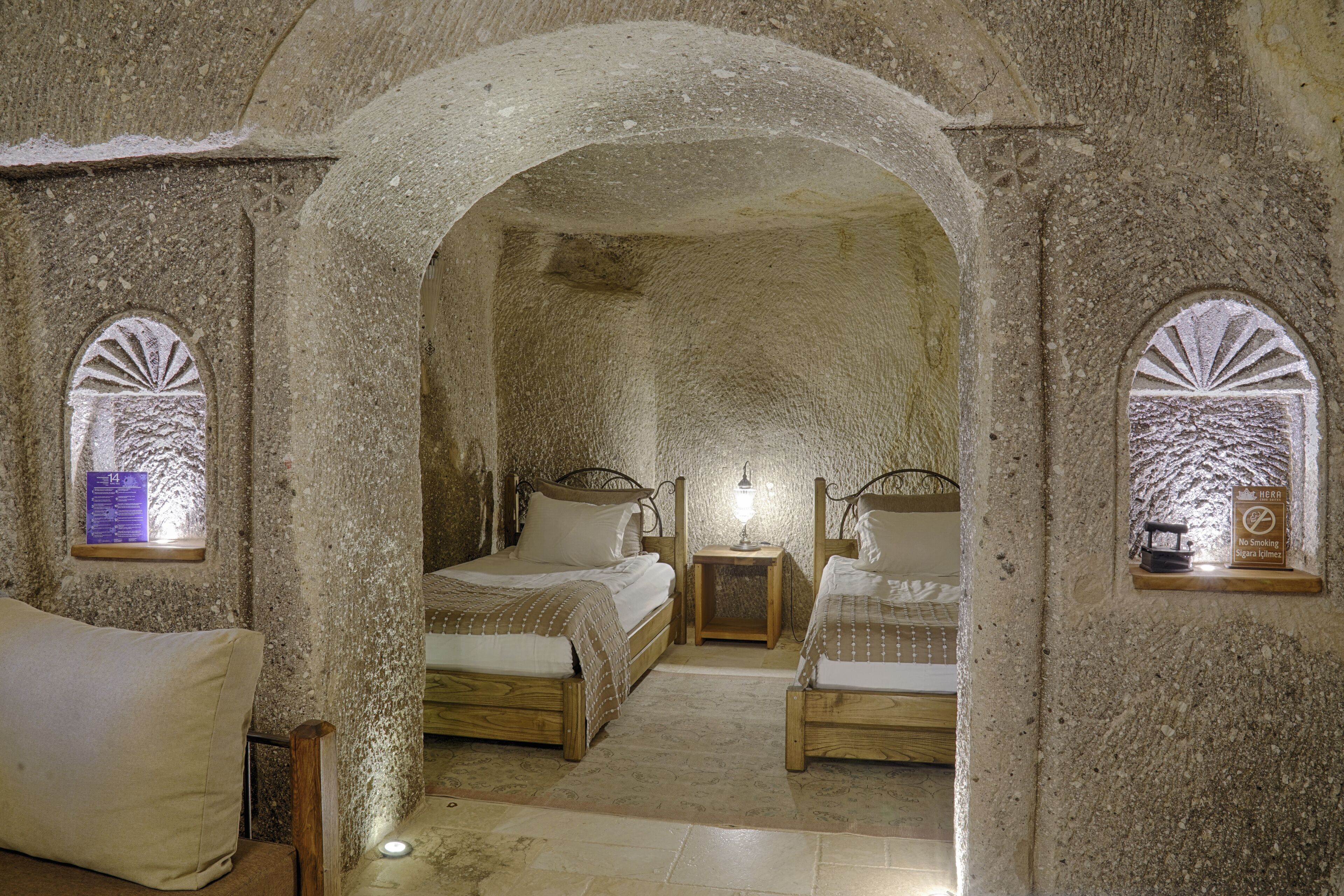 A stone-walled room with two wooden beds and a bench, featuring arched doorways and small windows with decorative patterns.