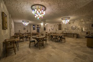 Un comedor amplio con varias mesas y sillas, candelabros ornamentados, paredes de piedra y un ambiente rústico.