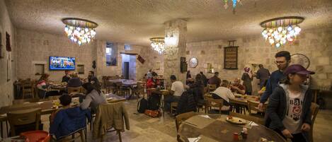 Un restaurante con varias mesas y sillas, una pantalla de televisión y lámparas colgantes de colores.