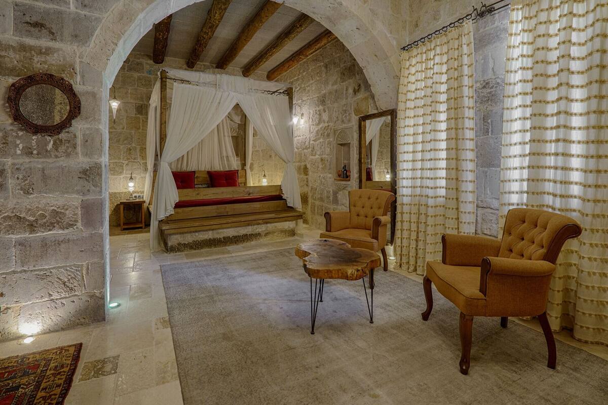 A stone-walled room with a four-poster bed, a wooden bench, and two armchairs.