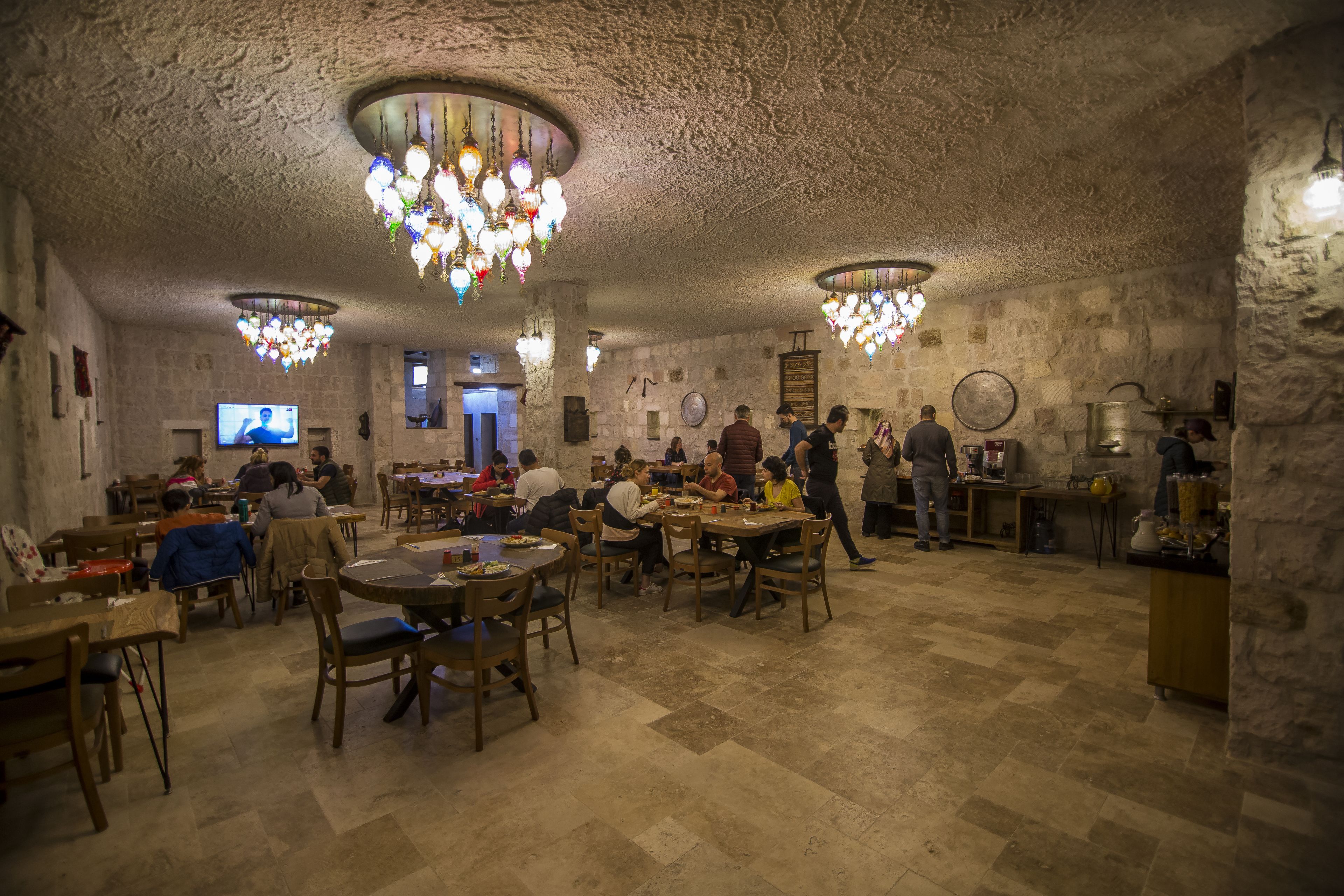 A dining area with stone walls and wooden tables, featuring chandeliers and a television screen.