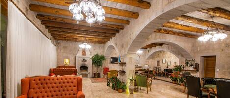 A spacious room with a fireplace, wooden beams, and a chandelier. There are multiple seating areas with chairs and a sofa.