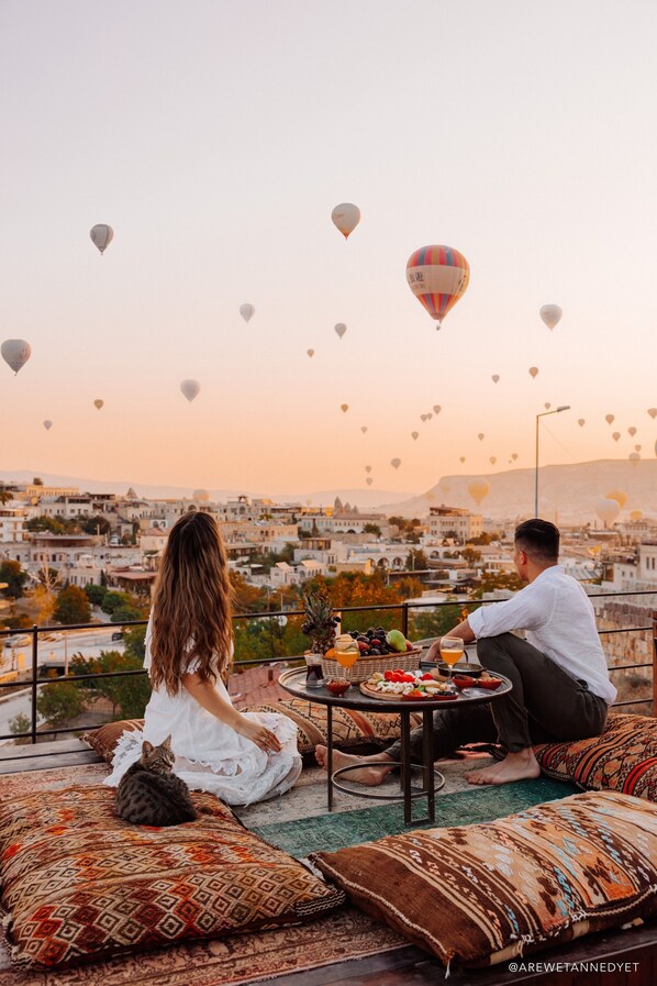 Una pareja sentada en un balcón con vistas a la ciudad, una mesa con comida y bebidas, y un gato.