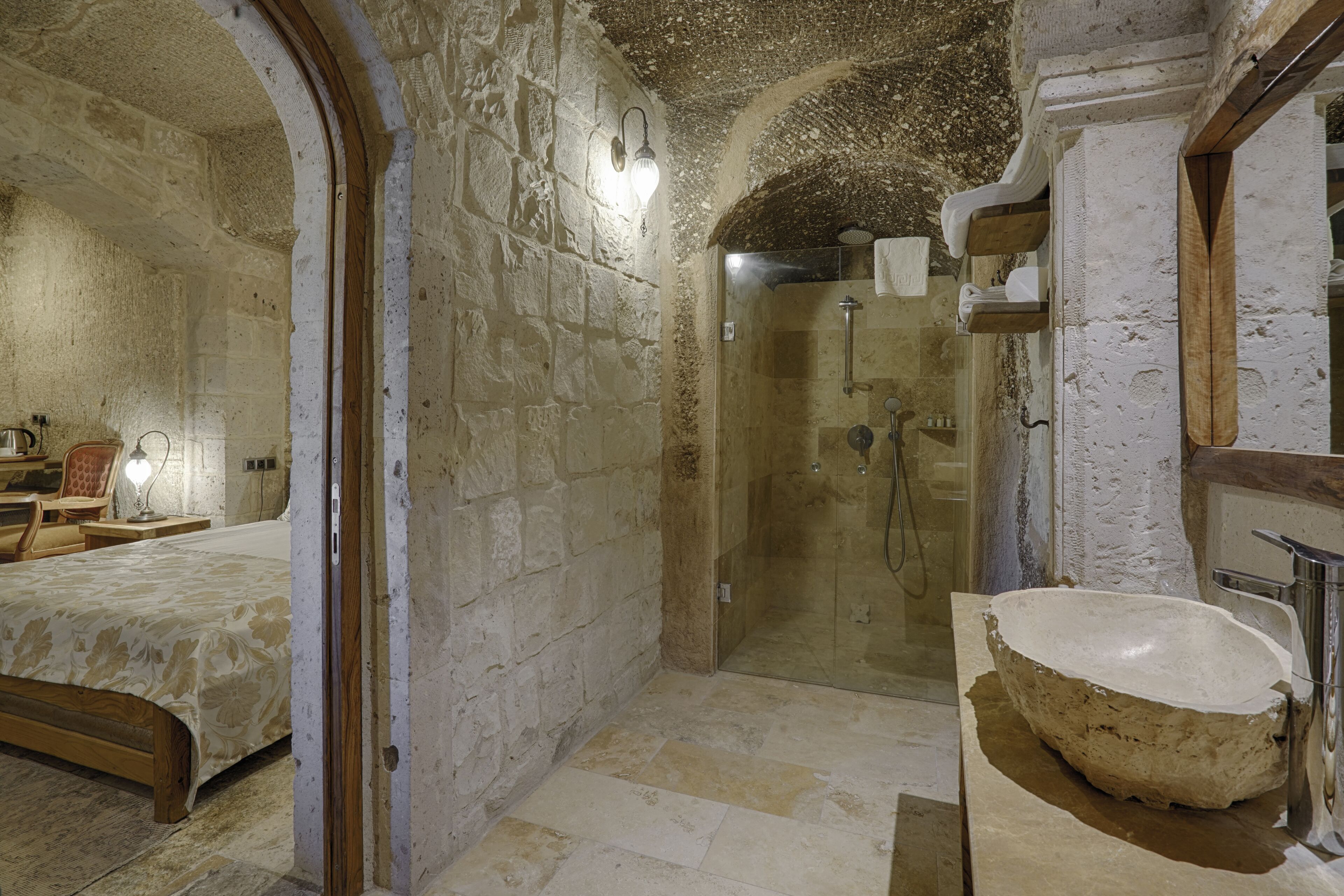 A stone-walled bathroom with a large basin, a shower, and a bed with a wooden headboard.