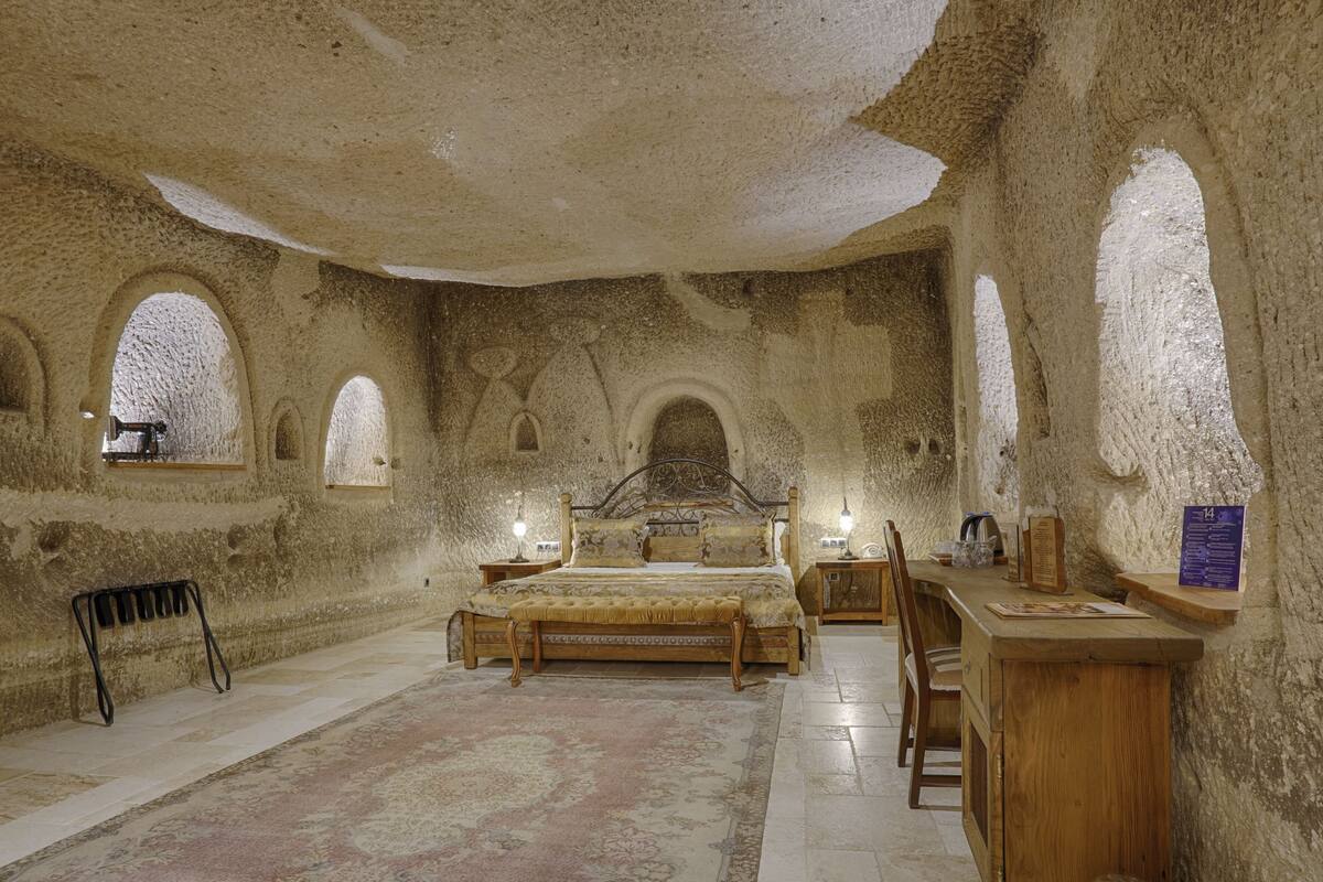 A room with a bed, a desk, and a chair. There are arched windows and a rug on the floor.
