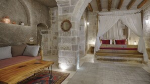 Una stanza rustica con pareti in pietra, una panca in legno, un letto con cuscini rossi e una struttura a baldacchino.