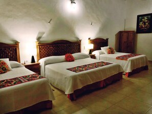 Ein Hotelzimmer mit drei Einzelbetten, jedes mit weißer Bettdecke und farbenfroher Decke. Es gibt ein Holz-Kopfteil, einen Nachttisch mit Lampe und einen Holzschrank.