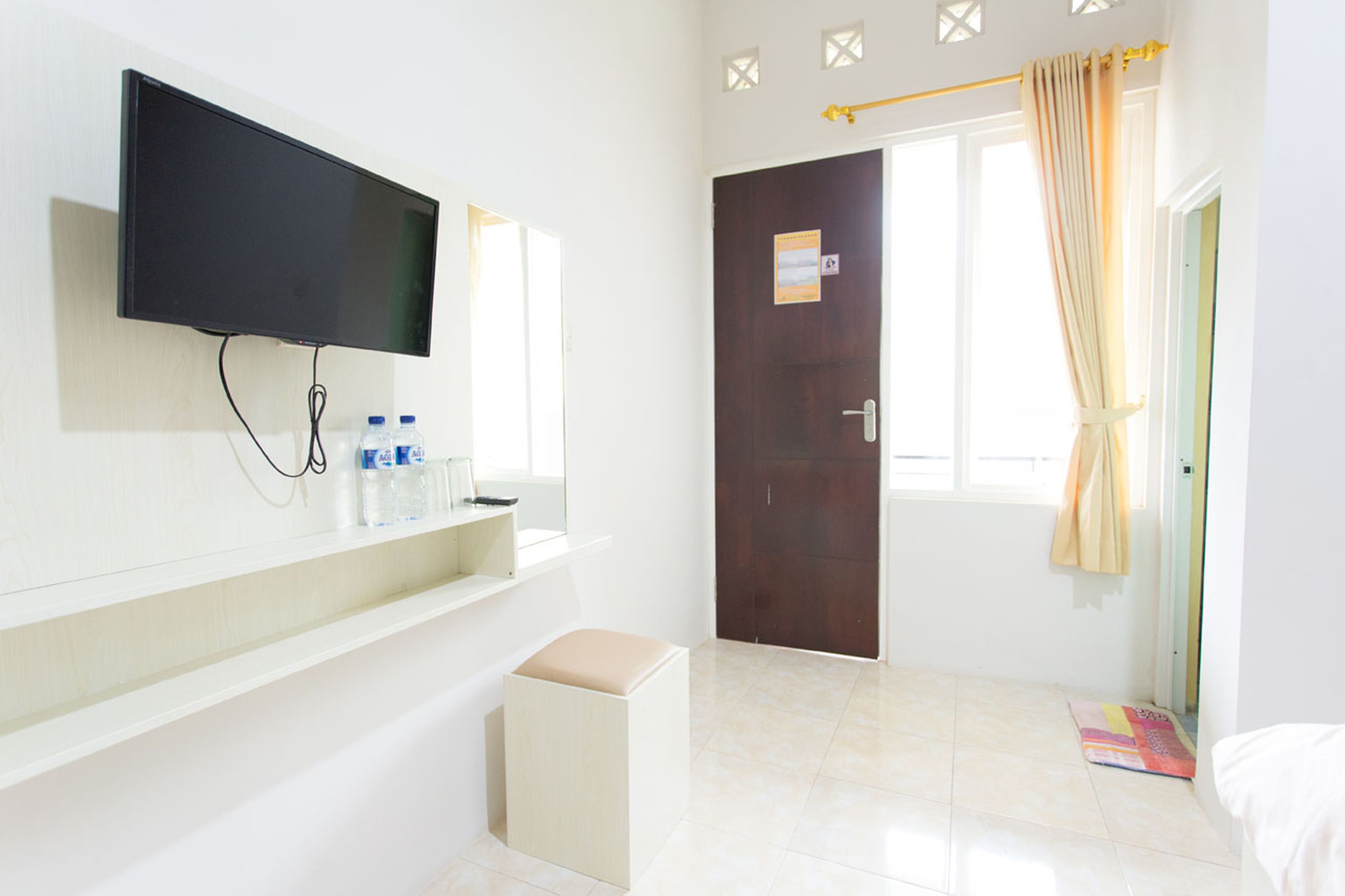 Habitación de hotel con una televisión de pantalla plana en la pared, un taburete pequeño, un estante con artículos de aseo, una puerta con un cartel y una ventana con cortinas.