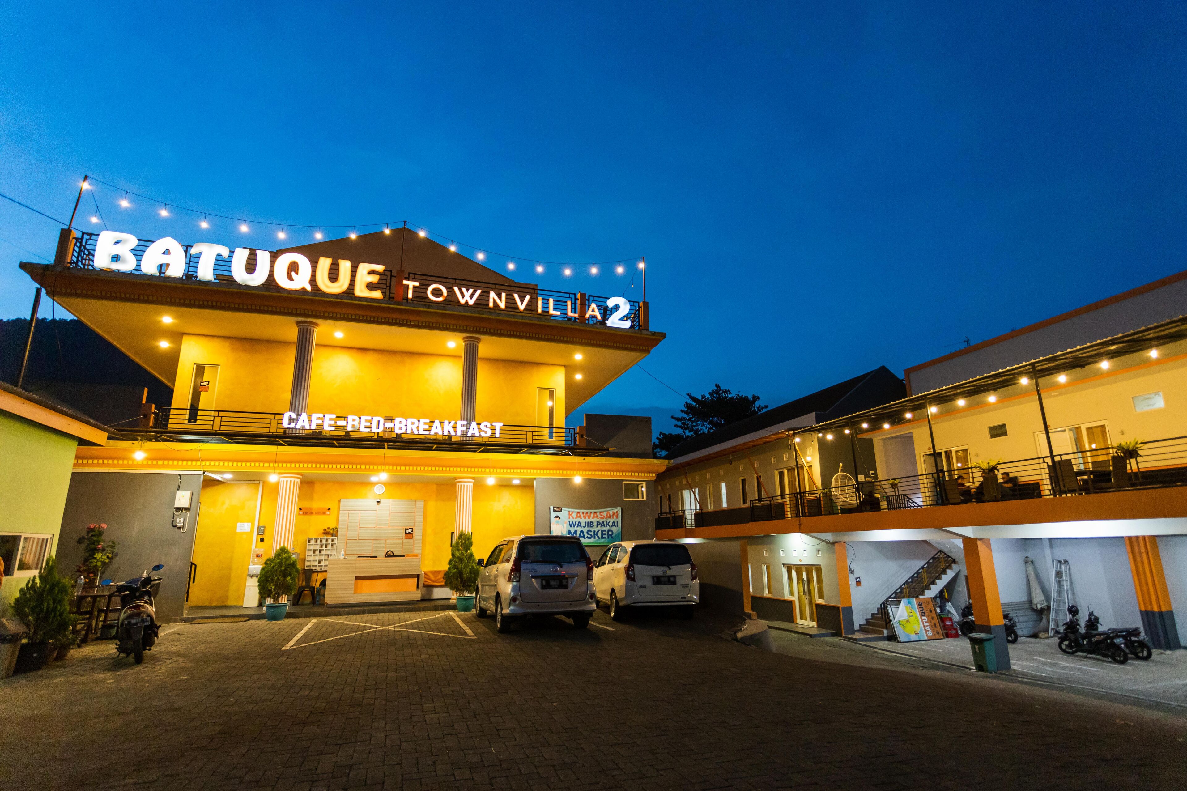 A building with a sign reading "Batuque Townvilla 2" and "Cafe-Bed-Breakfast". There are cars parked in front and a staircase leading to an upper level.