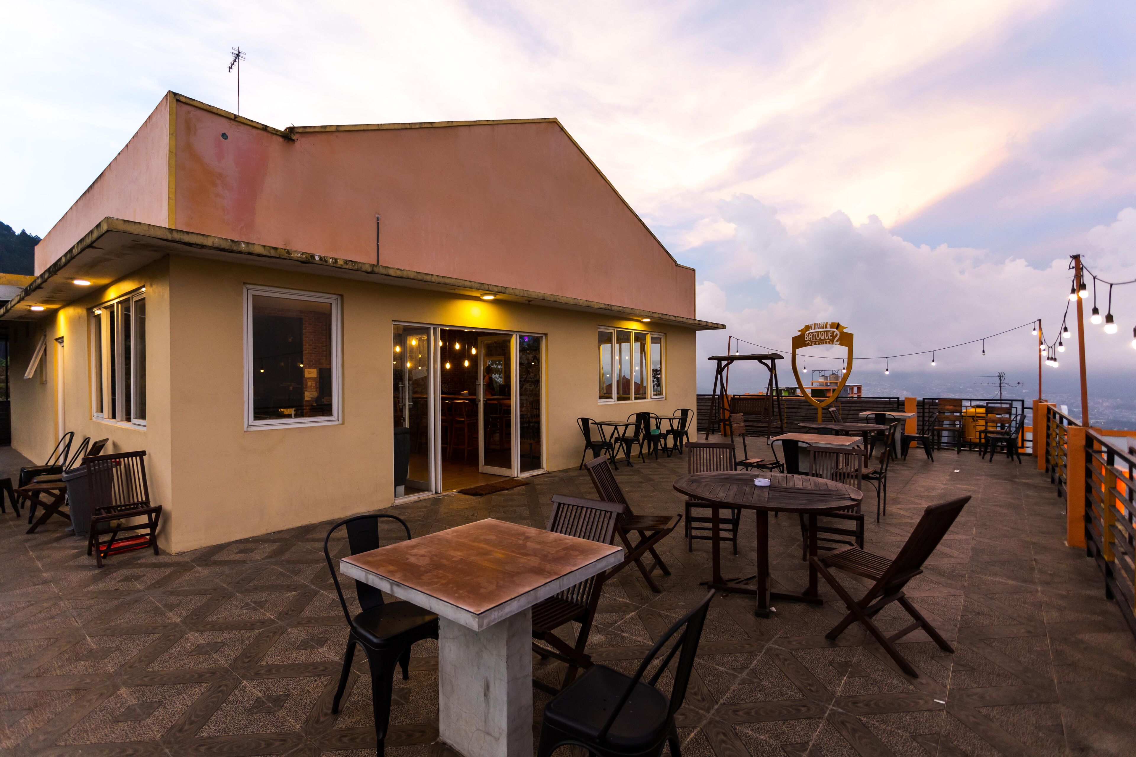 Uma área de terraço ao ar livre com mesas e cadeiras, um edifício com grandes janelas e uma placa que diz "Lounge Bar".