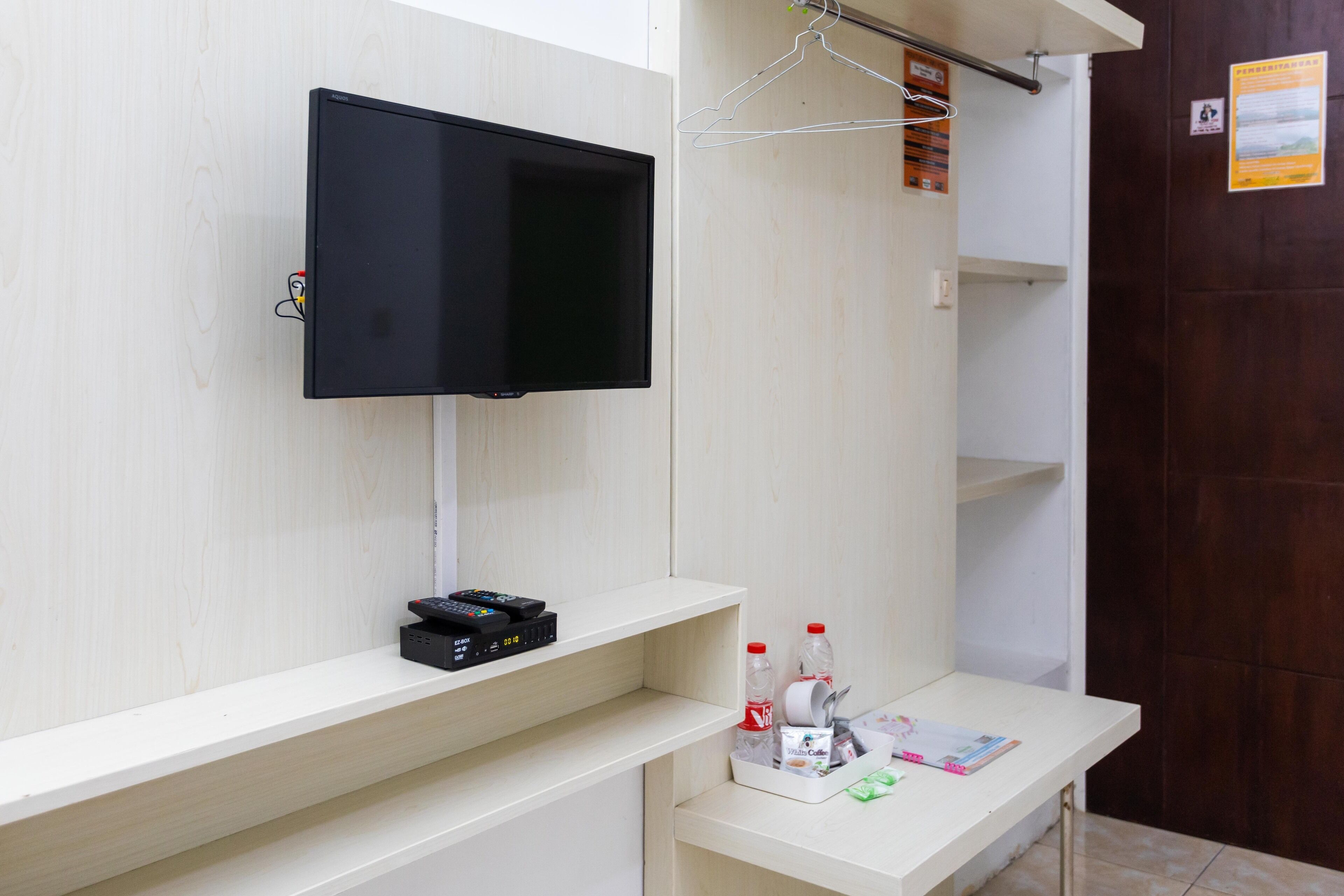 A hotel room with a mounted TV, a small control panel, and a shelf with toiletries.