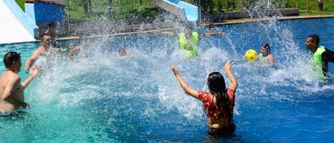 A swimming pool with people playing and splashing water, a slide, and a grassy area in the background.