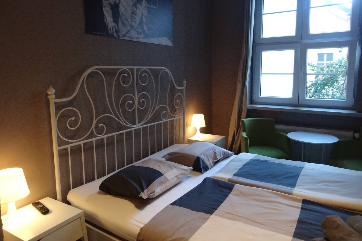 A neatly made bed with a quilt, a nightstand with a lamp and remote, a window with a view, and a green chair.