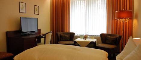 A hotel room with a bed, a desk with a television, two chairs, a small table with a vase, and curtains.