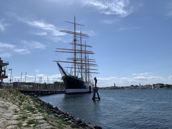 Et højt skib ligger ved et ankomsthavn med en klippefyldt kystlinje og en bro i baggrunden.