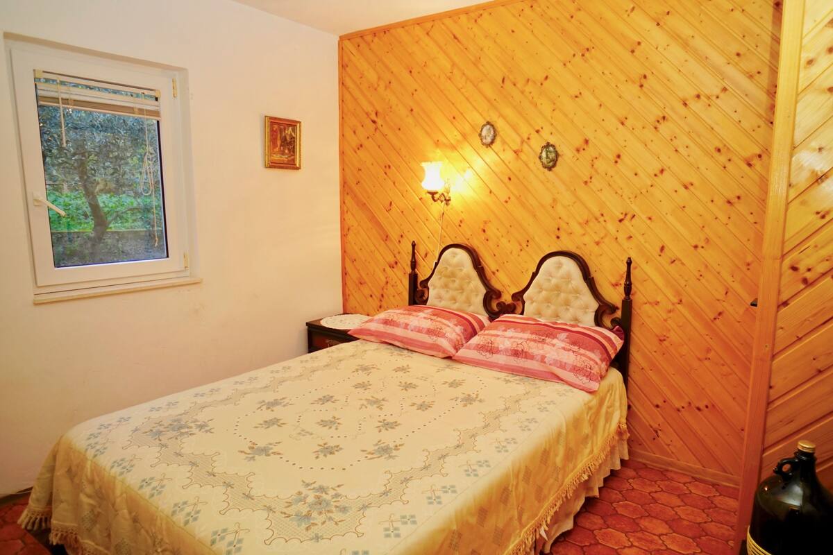 A bedroom with a wooden headboard, a bed with floral bedding, a nightstand, a window with a view of trees, and a wall-mounted light fixture.