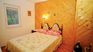 Un dormitorio con cabecera de madera, cama con ropa de cama floral, mesita de noche, ventana con vistas a árboles y una lámpara fijada a la pared.