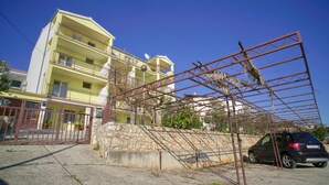 A multi-story yellow building with balconies, a metal structure with vines, and a parked car. - Sunset apartments (Seget)