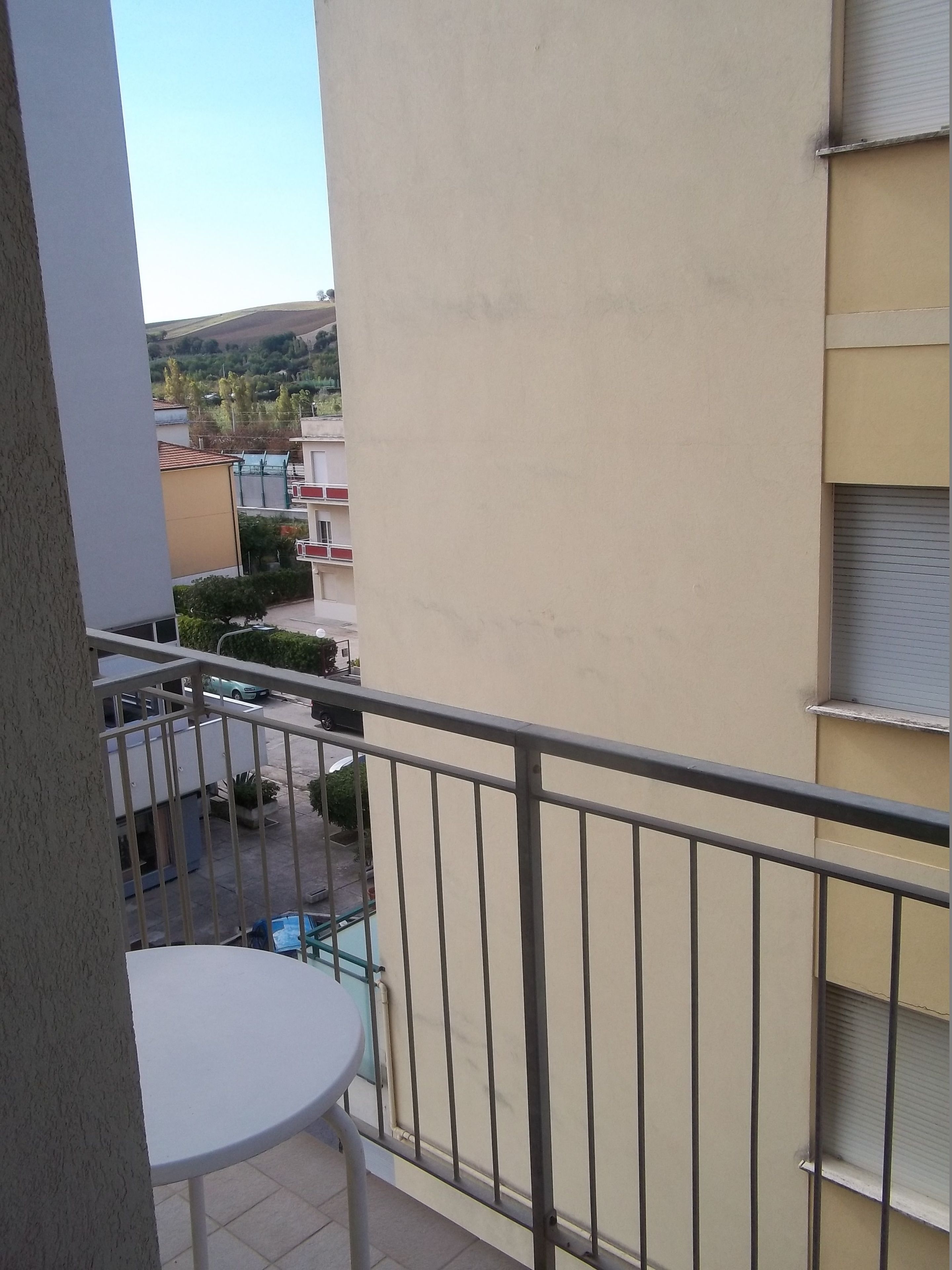 Un balcón con una mesa blanca y barandilla de metal, con vistas a una zona residencial con casas y vegetación.